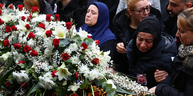 Mehrere Frauen stehen am Sarg von Ferhat Unvar in Hanau. Er ist eines der Opfer des rechten Anschlags. Die Frauen weinen oder blicken ernst. Auf dem Sarg liegt ein großes Bouqet aus Blumen.