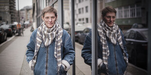 Eine junge Frau, kurze Haare, warme Jacke, steht draußen und spiegelt sich in einem Fenster.