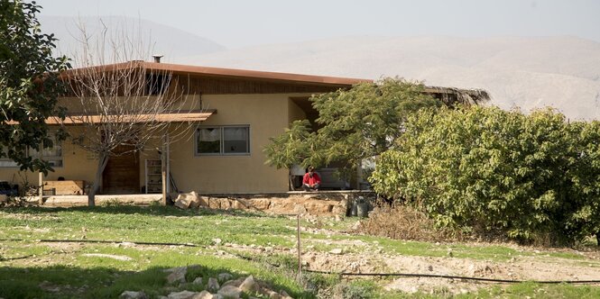 ein Haus mi Bäumen und Büschen daneben und davor. Auf der Terrasse sitzt ein Mann im Schneiderseitz