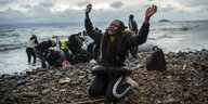 eine Frau kniet an einem Strand und hebt die Arme, hinter ihr Menschen, die ein Boot aus dem Wasser ziehen