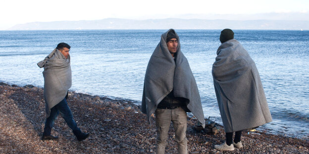 Migranten stehen in Decken gehüllt an einem Strand auf Lesbos.
