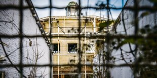 Gelbes halbrundes Gebäude am Ende einer Gasse durch ein Gitter gesehen, mitten auf dem Dach ein laternenförmiges Oberlicht