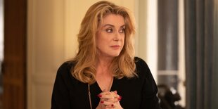 Catherine Deneuve steht in einem Zimmer, die Hände ineinander verschränkt, den Blick skeptisch zur Seite