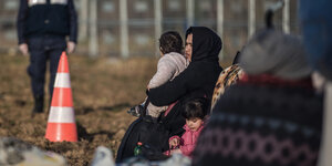 Flüchtlinge an der Grenze zwischen der Türkei und Griechenland: Eine Frau mit zwei kleinen Kindern wartet. Im Hintergrund sind ein Grenzzaun und ein griechischer Polizist zu sehen