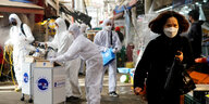 Straßenszene in Seoul: Eine Frau mit Schutzmaske, im Hintergrund Mitarbeiter einer Desinfektionsfirma in Arbeitskleidung
