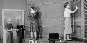 Frauen in den 1950er oder 60er Jahren schreiben mit Kreide Zahlen an Tafeln