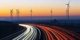 Lichter von Fahrzeugen auf einer Autobahn im Sonnenuntergang, im Hintergrund Windräder.