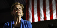 Elizabeth Warren, eine blonde Frau mit Brille, vor einer US-Fahne