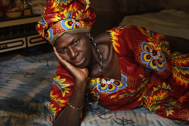 Eine Frau stützt ihre Hände unter dem Kinn auf, sie liegt mit bunten Kleidern und Kopfbedeckung auf einem Bett