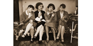 Vier junge Frauen sitzen nebeneinander auf einer Chaiselongue, eine Hand jeweils aufs Herz gelegt, die Köpfe leicht geneigt.