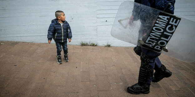 ein kleiner Junge in Winterjacke, an dem ein Polizist mit Schutzschild vorbeiläuft. Dieser ist nur von den Oberarmen abwärts zu sehen