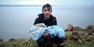 Ein erschöpfter Mann mit nassen Haaren steht an einer Steilküste und hält ein Baby im Arm