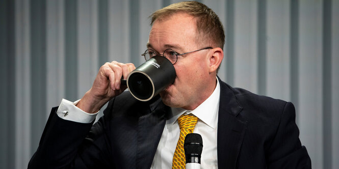 Mulvaney hält eine schwarze Kaffeetasse an Mund und Nase, er sitzt vor einem grauen Vorhang