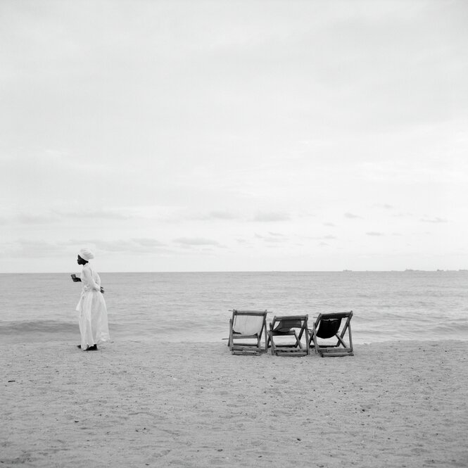 Ein Mann läuft am Strand entlang mit weißem Gewand, es stehen drei Strandliegen verlassen hinter ihm, vor ihm das Meer