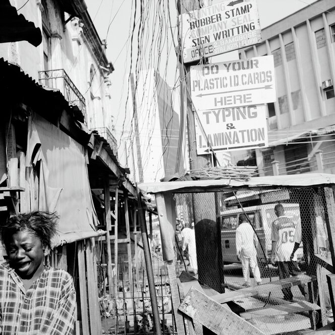 Eine Straßenszene in Lagos, ein Mädchen steht weinend im Vordergrund, dahinter ein Schild mit der Schrift: Do your Plastic Cards here typing and lamination, schwarz-weiß Foto