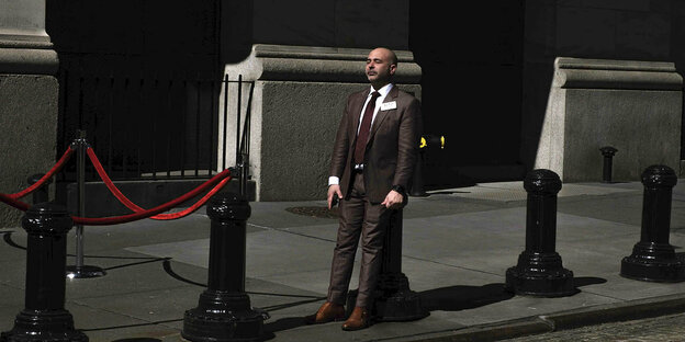 Ein Mann in braunem Anzug ruht sich auf einem Poller in der Wall Street in New York aus