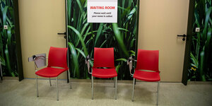 rote Stühle vor zwei geschlossenen Türen, ein Schild an der Wand, darauf steht in deutsch und englisch: Wartebereich. Bitte warten Sie, bis Sie aufgerufen werden.