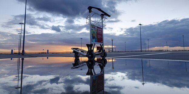 leerer Parkplatz am Flughafen Köln Bonn, in einer Pfütze spiegelt sich der Himmel, in der Bildmitte steht ein Kofferkuli