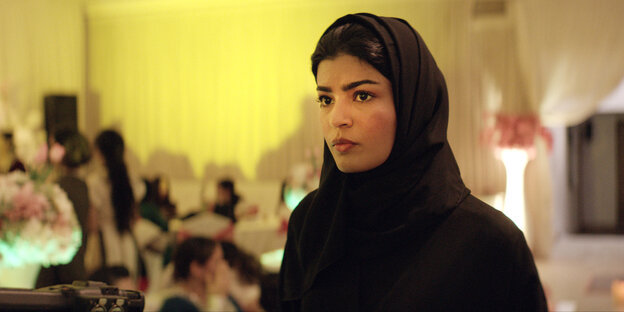 Eine junge Frau mit Niqap in einem Innenraum, weitere Personen nur schemenhaft zu erkennen.