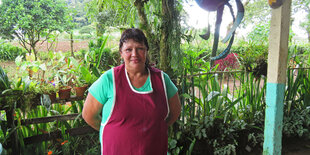 Maribel in roter Schürze steht auf einer Veranda mit Blick in den Nutzgarten