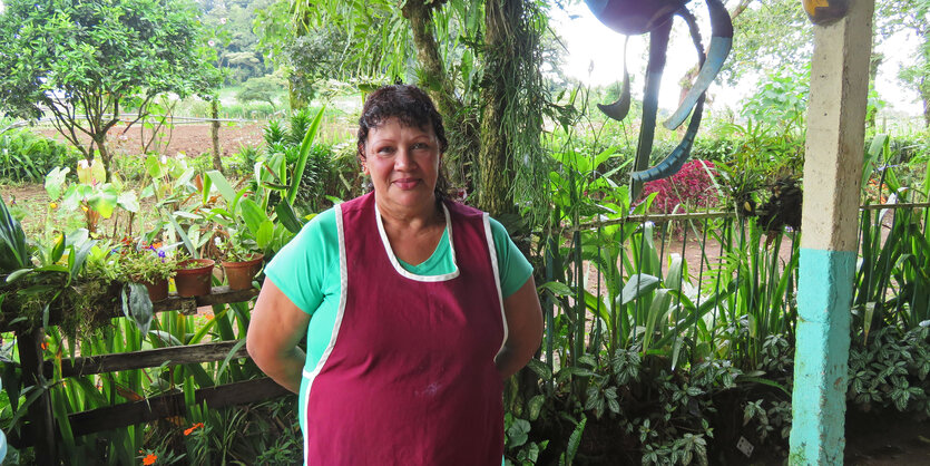 Maribel in roter Schürze steht auf einer Veranda mit Blick in den Nutzgarten