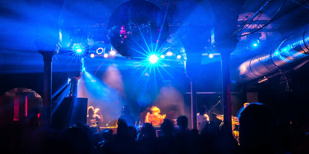 Ein Club ist in blaues Licht getaucht, im Vordergrund tanzen Menschen auf der Bühne wird Musik gemacht