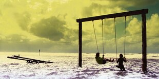 Zwei Kinder auf einer überspülten Schaukel auf einem Strand.