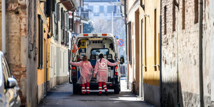 Eine Straße, man sieht einen Krankenwagen und zwei Pflegekräfte von hinten die Straße entlang fahren