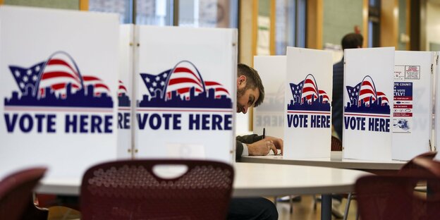 Ein Mann sitzt in einem Wahllokal und wählt. Er hat kurze dunkle Haare. Die Abtrennungen zwischen den Wahlkabinen sind beschriftet: "Vote here"