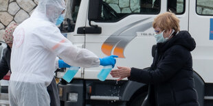 ein Mann mit Schutzanzug und -maske desinfiziert einer Frau mit Mundschutz die Hände