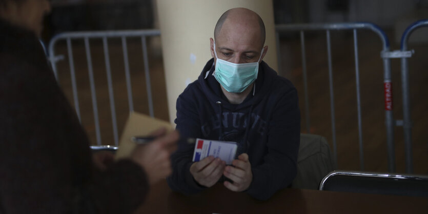 Ein Mann mit Mundschutz sitzt hinter einem Tisch und hält ein Stück Papier in der Hand