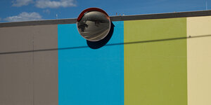 Eine dreifarbige Wand an der oben ein runder Spiegel hängt, darin spiegelt sich eine leere und grpße Straße