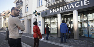 Fünf Menschen stehen mit großem Sicherheitsabstand zueinander vor einer Apotheke und warten darauf, dass sie reingelassen werden
