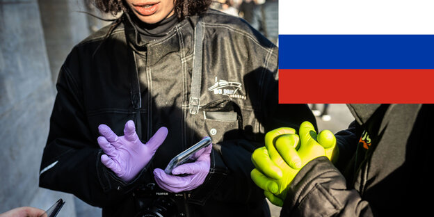Eine Frau mit lila Plastikhandschuhen hat ein Handy in der Hand. Oben rechts ist eine russische Flagge abgebildet und über das Foto drübergelegt