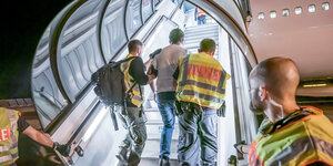 Zwei Polizisten in gelben Warnwesten führen einen Mann eine Treppe hoch, die in ein Flugzeug führt