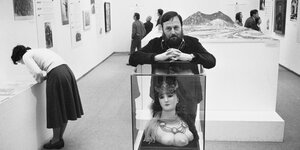 Schwarz-weiß-Foto von Harald Szeemann, der sich auf eine Vitrine in einer Ausstellungshalle stützt. In der Vitrine eine Frauenbüste mit drei Brüsten