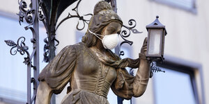 Die Skulptur "Die Schneidersfrau" auf dem Heinzelmännchenbrunnen in der Kölner Altstadt hat eine Atemschutzmaske um.