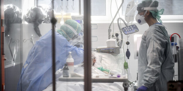 Menschen in medizinischer Schutzkleidung und Atemmasken behandeln hinter Glas einen Patienten.