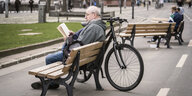Ein Mann sitzt auf einer Parkband, die auf einer Straße steht und liest in einem Buch.