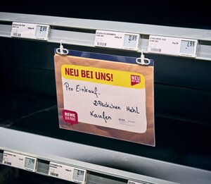 In einem leeren Supermarktregal hängt ein Schild: Pro Einkauf. 2 Päckchen Mehl kaufen In einem leeren Supermarktregal hängt ein Schild: Pro Einkauf. 2 Päckchen Mehl kaufen