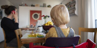 Eine Frau im Homeoffice an ihrem Laptop – sie telefoniert, während ihr Kind neben ihr in einem Kinderstuhl am Tisch sitzt