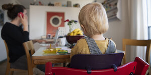 Ein Kleinkind von hinten, es sitzt an einem Küchentisch, im Hintergrund eine Frau vor einem Laptop