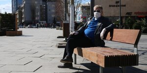 Ein Mann mit Mundschutz sitzt in der Sonne auf einer Bank.