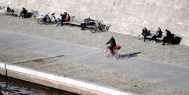 Menschen am Spreeufer sitzen auf Bänken, eine Person fährt auf dem Rad vorbei