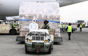 Zwei Männer, einer mit Schutzanzug, einer mit Sonnenhut, transportieren mit einem Wagen auf einem Flughafen Pakete.