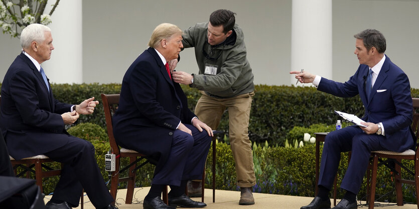 Donald Trump und Mike Pence sitzen auf Stühlen im Freien, ein Maskenbildner arbeitet am Präsidenten.