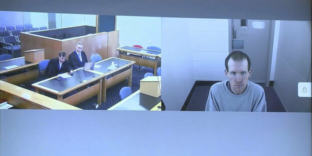 In der linken Bildhälfte sieht man einen fast leeren Gerichtssaal, rechts den per Video zugeschalteten Angeklagten