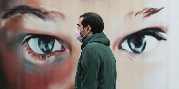 Ein Mann mit Mundschutz läuft vor einem Plakat mit riesigen Augen vorbei.