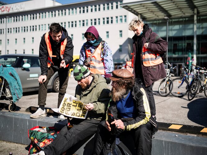 Drei junge Menschen in Warnwesten sprechen zwei Männer mit Bierflaschen vor einem Bahnhof an