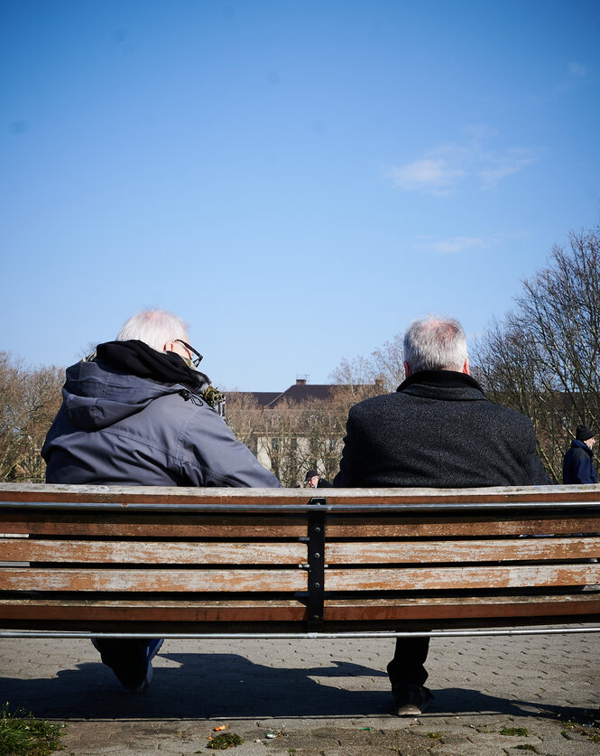 Corona in Berlin: Zwei Männer sind von hinten zu sehen, wie sie bei schönem Wetter auf einer Parkbank sitzen